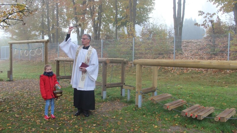 Pfarrer Konrad segnet den neuen Bewegungsparcours der Grundschule Viechtach und auch die umstehenden Gäste - Schülerin Annika Schiller hält den Weihwasserkessel.