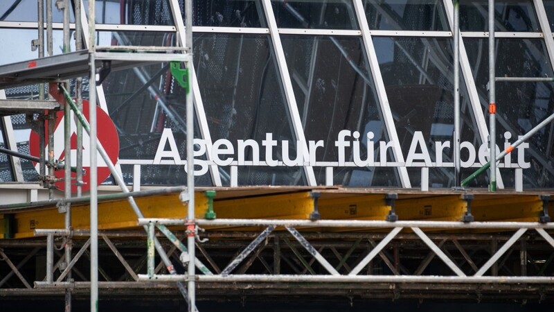 Der Schriftzug "Agentur für Arbeit" und das Logo der Behörde befinden sich hinter einem Baugerüst. Die Arbeitslosigkeit in Bayern ist fast unverändert.
