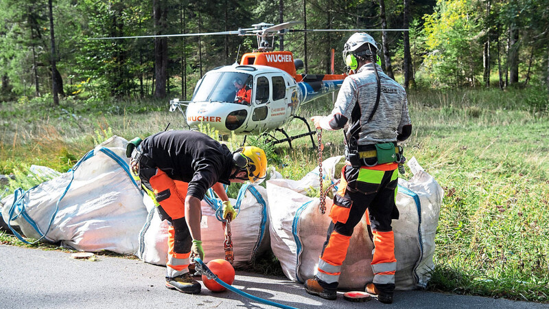 Besatzungsmitglieder eines Hubschraubers binden auf einer Waldlichtung zwei Säcke mit circa 1 240 Tannen- und Lärchensetzlingen an einen Hubschrauber. Gleich hebt er ab in Richtung des Gipfels des Rotkogels.