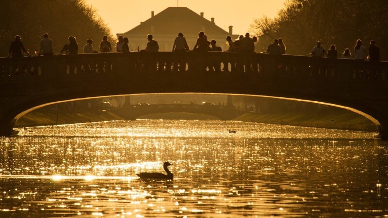 Menschen genießen den warmen Frühlingstag im goldenen Licht der untergehenden Sonne am Nymphenburger Kanal, der zum Schloss Nymphenburg (im Hintergrund) führt.