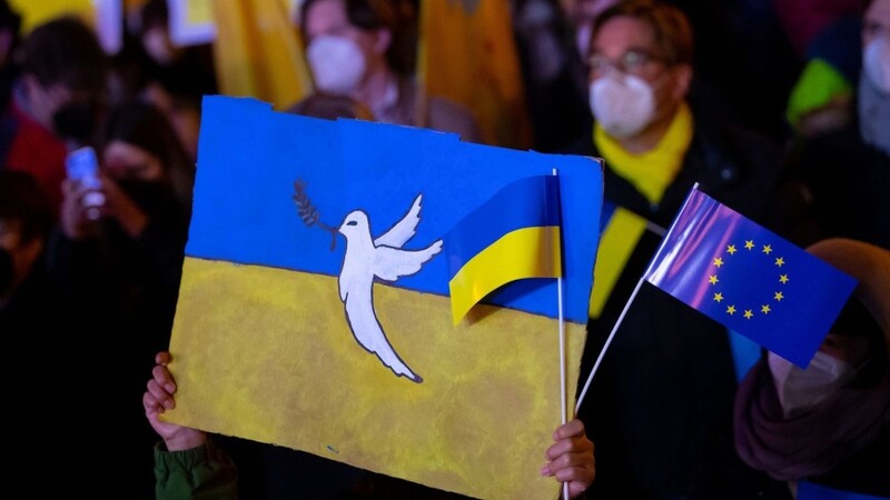 In München hält ein Kind bei einer Veranstaltung Schilder mit einer Friedenstaube und der Flagge der Ukraine in die Höhe. (Archivbild)