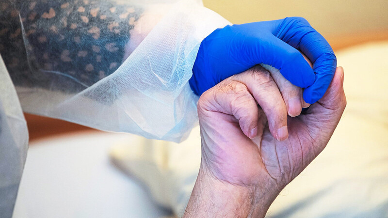 Eine Altenpflegerin in Schutzausrüstung hält die Hand eines Bewohners. Immer wieder kommt es in Seniorenheimen zu Coronavirus-Ausbrüchen, auch in der Region gab es bereits einige.