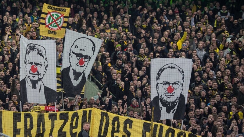 BVB-Fans zeigen Banner mit den Gesichtern von Dietmar Hopp, Fritz Keller und Karl-Heinz Rummenigge (v.l.) mit roter Clownsnase. Darunter ist ein Banner mit der Aufschrift "Die hässlichen Fratzen des Fußball".