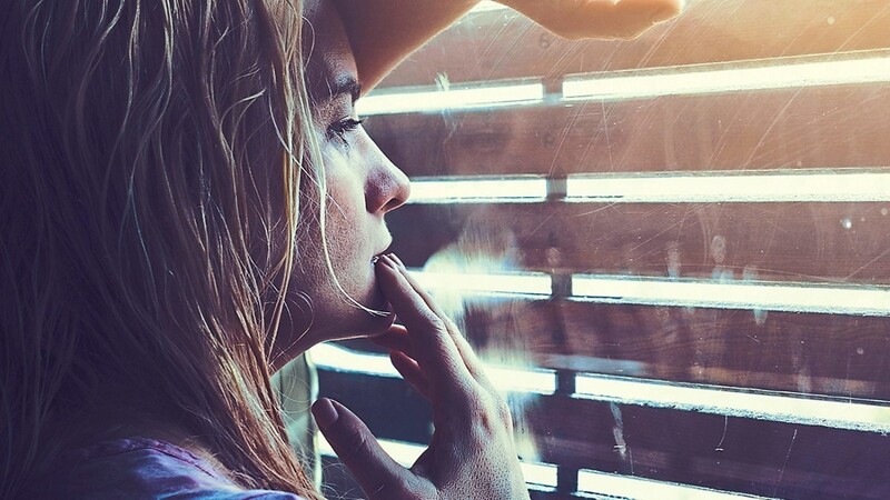 Das Symbolfoto zeigt eine junge Frau an einem abgedunkelten Fenster. Besonders Jugendliche treffen die Corona-Beschränkungen hart.
