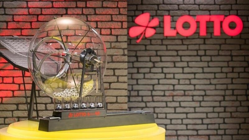 Eine Ziehungsmaschine der Lottozahlen 6 aus 49 steht neben dem "Lotto"-Logo.