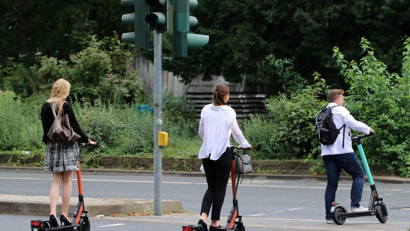 Ein Mann und zwei junge Frauen überqueren mit ihren E-Scootern an einer Ampel die Straße. Versicherer fordern aber aufgrund der vielen Unfälle eine Helmpflicht.