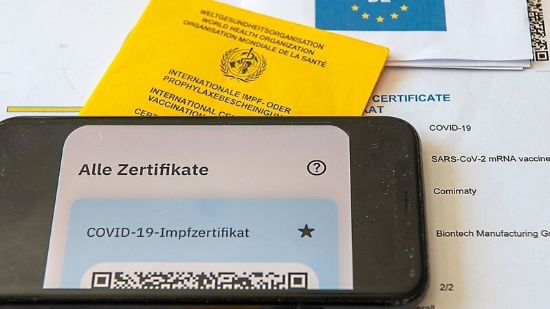 Ein Impfpass und ein Smartphone, auf dem die App CovPass läuft, liegen auf einem Impfzertifikat, das von einer Apotheke ausgestellt wurde. In bayerischen Apotheken versuchen immer wieder Menschen mit Wohnsitz in den Nachbarländern, mit gefälschten Impfpässen digitale Zertifikate zu bekommen.(Symbolbild)