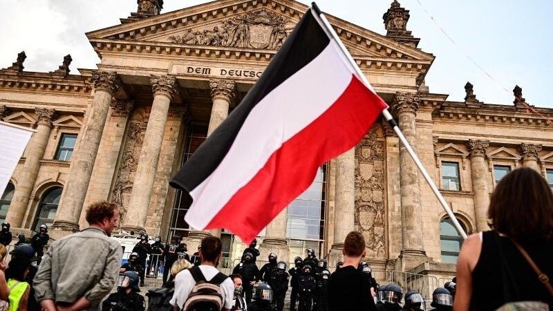 Teilnehmer einer Kundgebung gegen die Corona-Maßnahmen stehen vor dem Reichstag, ein Teilnehmer hält eine Reichsflagge mit schwarz-weiß-roten Querstreifen.