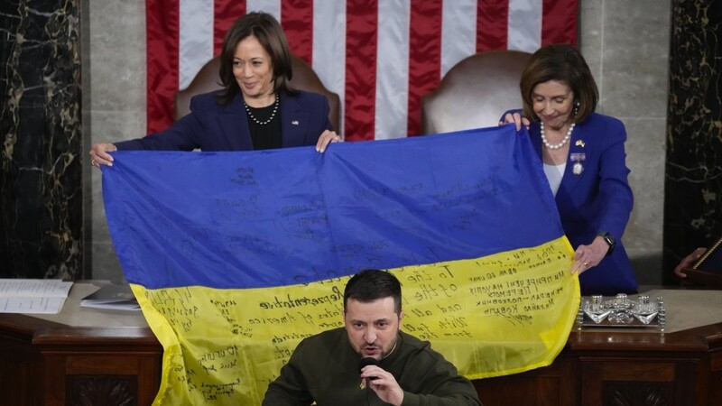 Wolodymyr Selenskyj, Präsident der Ukraine,, spricht vor einer ukrainischen Flagge, die von Kamala Harris (l.), Vizepräsidentin der USA, und Nancy Pelosi, Sprecherin des Repräsentantenhauses, gehalten wird.