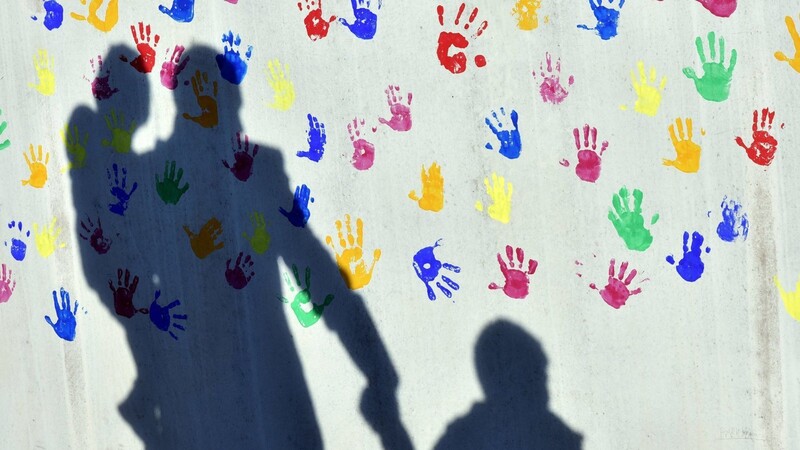 Der Schatten von einem Mann mit zwei Kindern zeichnet sich an der Wand einer Kindertagesstätte ab, die mit bunten Handabdrücken verziert ist.