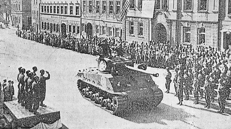 Amerikanische Mannschaften, eine Musikkapelle und Panzer paradieren vor zahlreichen Zuschauern am 18. August 1945 auf dem Vilsbiburger Stadtplatz. Rechts das von den Amerikanern als Militärregierung mit dem Sternenbanner besetzte Finanzamt-Gebäude.