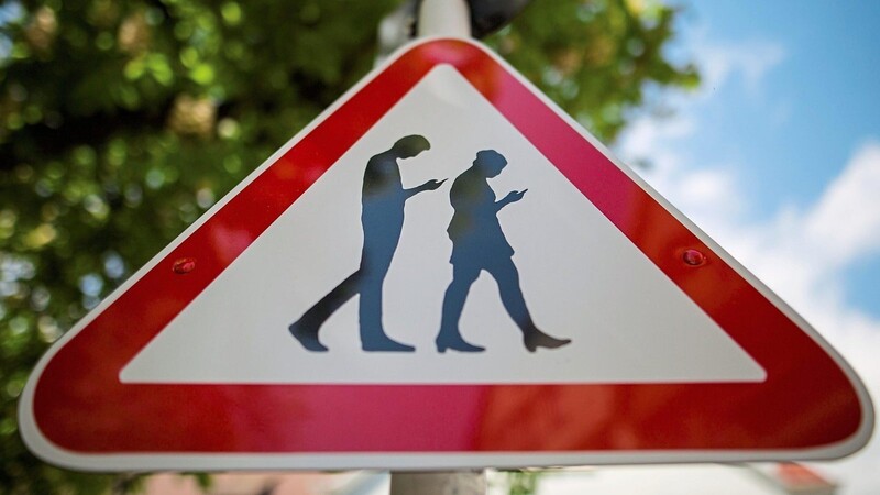 Ein Schild warnt an einer Straße neben einer Schule vor "Smombies". Die Abkürzung ist ein Kunstwort für Smartphone und Zombie und symbolisiert Menschen, die ständig mit dem Blick auf das Smartphone über die Straßen laufen.