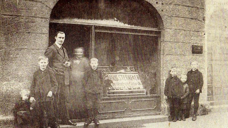 Das Foto aus dem Jahre 1924 zeigt den Tabakfabrikanten Ludwig Sternecker in der Aprilgasse 5 in der Ladentür mit seiner Frau, die sich wahrscheinlich nicht so still gehalten hat, wie es der Fotograf forderte. Ihr Gesicht ist leider verwackelt. Links sind drei Kinder zu sehen, rechts stehen nochmals drei.