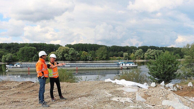 Dominik Zehatschek und Christian Eckl (v. l.) von Wiges im Gespräch über den neuen Querdeich bei Waltendorf (r.), der gerade mit Steinen beschwert und fertiggestellt wird. Während im Uferbereich der neue Hochwasserschutz gebaut wird, läuft die Schifffahrt auf der Donau weiter.