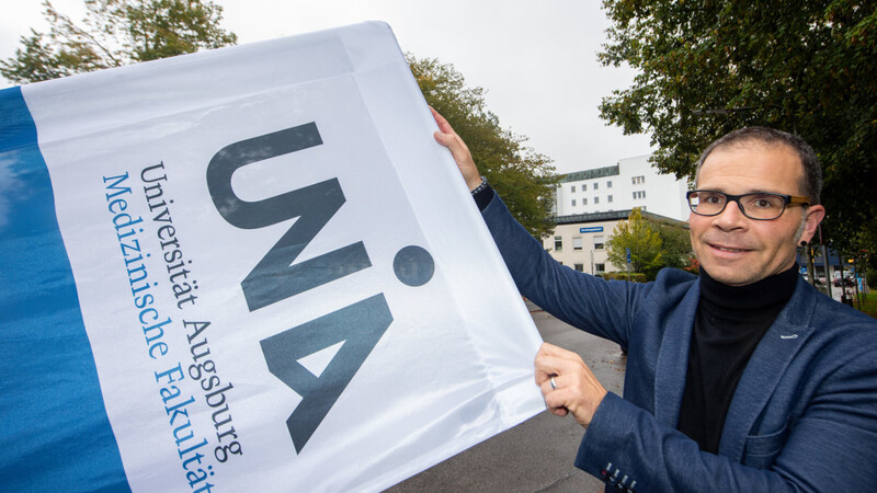 Jörn Böke, Geschäftsführer der medizinischen Fakultät der Universität Augsburg, hält vor dem Lehrgebäude ein Banner mit dem Logo und Schriftzug der Fakultät.
