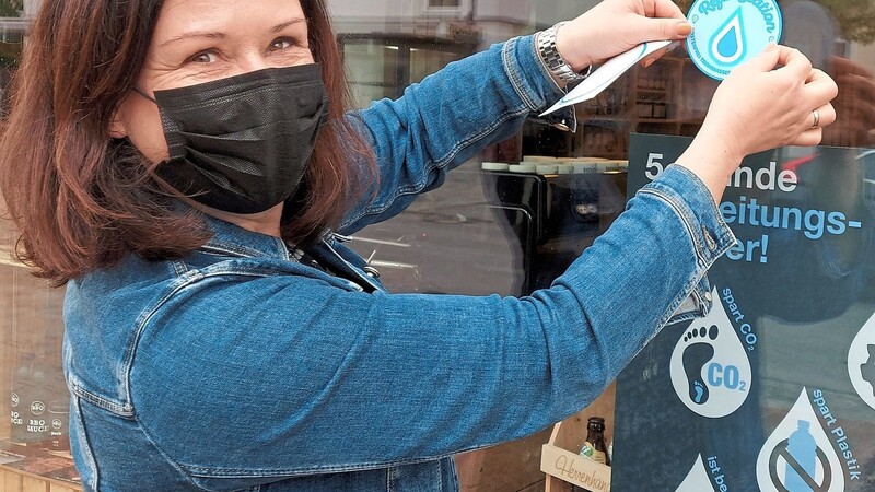 Monika Pernstecher, die Inhaberin des Puralei, bringt den Aufkleber von Refill Deutschland an ihrem Schaufenster an.