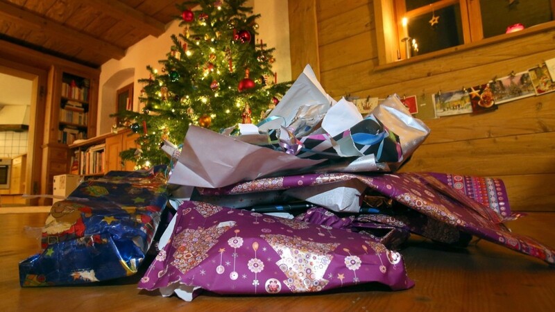 Zerknülltes weihnachtliches Geschenkpapier liegt vor einem geschmücktem Christbaum. (Symbolbild)