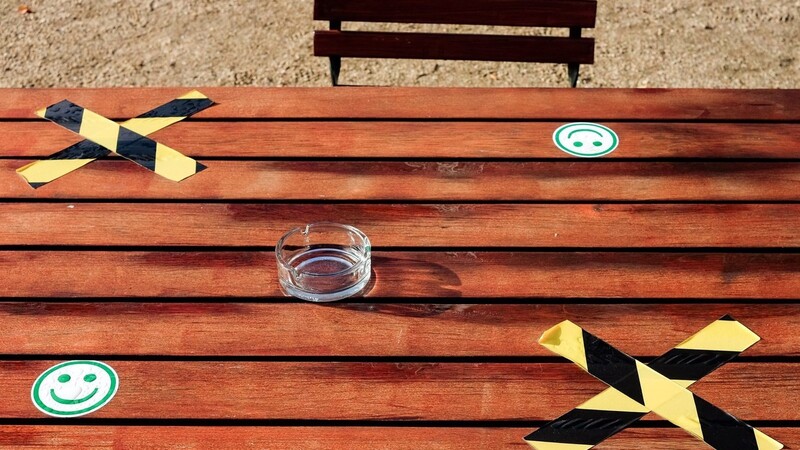 Auf einem Tisch eines Freiluftcafes steht zwischen aufgeklebten Kreuzen und Smileys für den Corona-Abstand ein leerer Aschenbecher.
