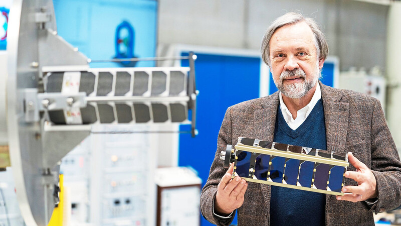 Der Präsident des Zentrums für Telematik, Klaus Schilling, steht vor einem Dynamiksimulator, an dem ein NetSat-Pico-Satellit angebracht ist und hält ein Modell eines solchen Kleinstsatelliten in den Händen.