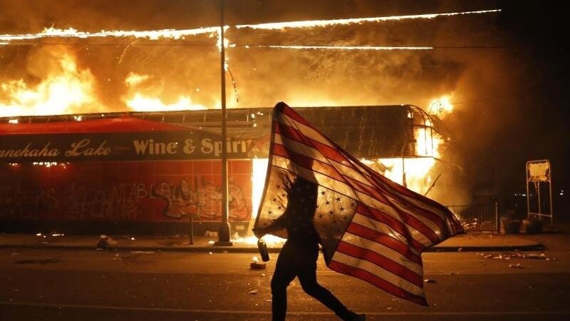 Ein Mann mit umgedrehter US-Flagge vor einem brennenden Gebäude in Minneapolis.