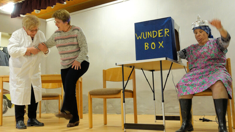 Der Wunderdoktor (Rita Pfeffer) kann mit seiner Wunderbox angeblich alle Krankheiten heilen, auch das Beinreißen (Bernadette Fischer) oder das Armzucken (Ilse Lorenz, rechts).