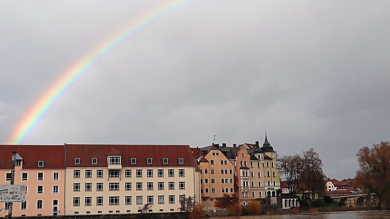 Grell leuchtende Farben durchbrechen das Regenwolkengrau bei der Weinlände an der Donau, wo sich ein Regenbogen von hinter den Häusern über das Wasser spannt.