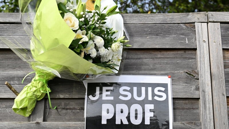 Ein Strauß Blumen hängt an einer Holzwand in der Nähe der Schule Bois d'Aulne in Conflans-Sainte-Honorine, neben einem Zettel mit der Aufschrift "Je suis Prof" (Ich bin Lehrer) zu Ehren von Samuel Paty. Sechs Jugendliche sitzen wegen etwaiger Beteiligung an seiner Enthauptung auf der Anklagebank.