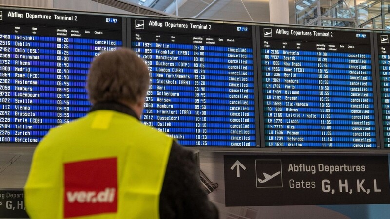 Ein Streikposten der Gewerkschaft verdi steht am Flughafen München vor einer Anzeigetafel.