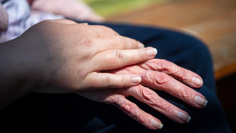 Die beschützende Hand einer Besucherin liegt auf der Hand einer Bewohnerin eines Seniorenheims.