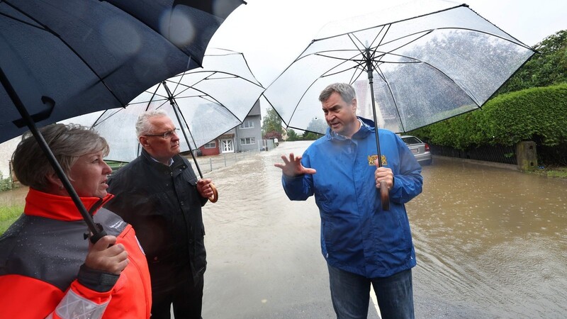 Ministerpräsident Markus Söder (r.) und sein Innenminister Joachim Herrmann stehen am Rande einer überfluteten Straße und unterhalten sich mit Helferin.