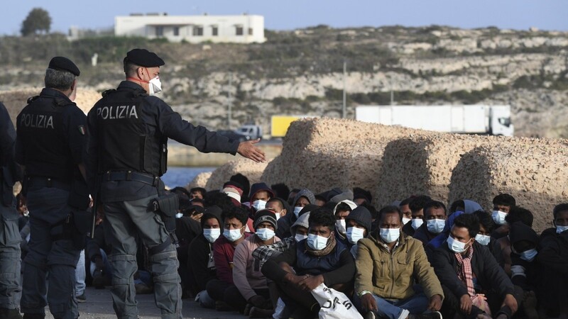 Italienische Polizisten (l.) stehen neben Migranten, die Mund-Nasen-Schutz tragen und auf dem Boden sitzen. Auf der kleinen italienischen Mittelmeerinsel Lampedusa kommen immer wieder Hunderte Bootsmigranten an.
