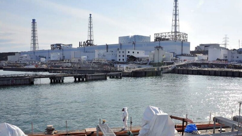 Ein Handout-Foto des japanischen Verteidigungsministeriums zeigt die Ruinen des havarierten Atomkraftwerks Fukushima von der Seeseite aus, während im Vodergrund eine Experte in Schutzkleidung zu sehen ist (Archivfoto vom 03.04.2011).