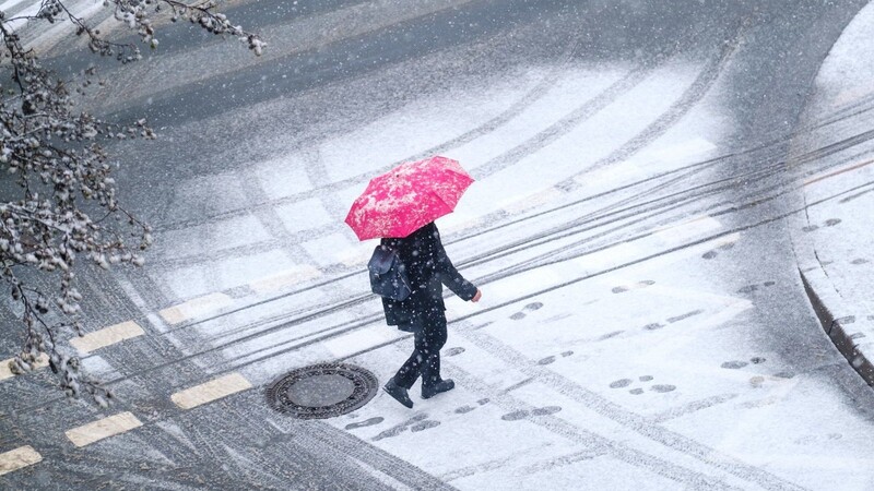Eine Frau geht mit einem Regenschirm über eine verschneite Straße in der Innenstadt. Der deutsche Wetterdienst warnt vor Schnee und Glätte in Bayern. (Symbolbild)