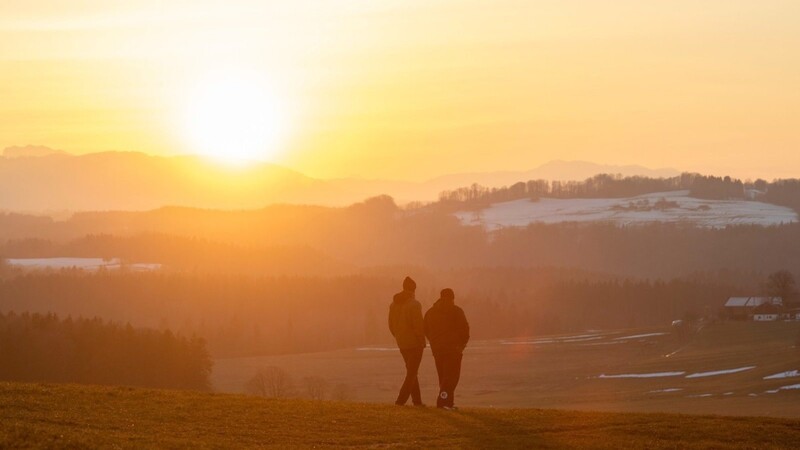Zwei Männer gehen bei Sonnenuntergang über eine Wiese auf dem Irschenberg. Das Wochenende in Bayern soll wieder sonnig werden.
