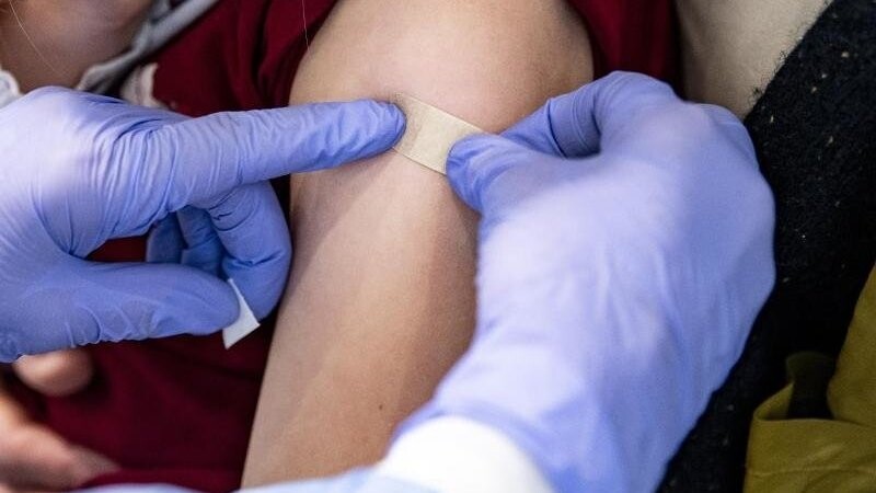 Eine Ärztin klebt einem 6-jährigen Mädchen, dass auf dem Schoß der Mutter sitzt, ein Pflaster auf die Impfstelle.