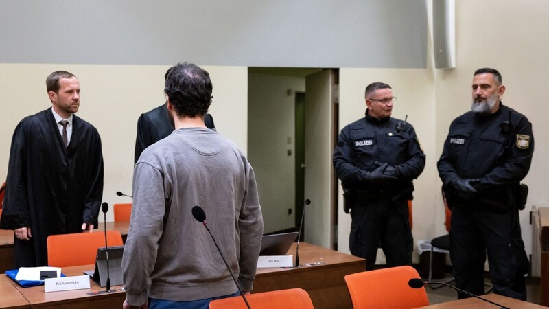 Der Angeklagte (3.v.l.) steht zusammen mit Polizeibeamten und seinen Anwälten Martin Gelbricht (l) und Maximilian Bär (2.v.l. verdeckt) im Verhandlungssaal.
