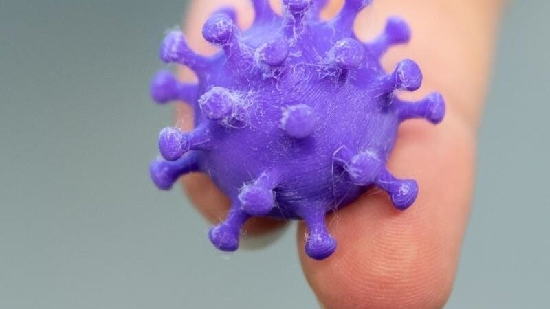 Eine Medizinisch-technische Assistentin hält auf zwei Fingern ein 3D Druck von einem Coronavirus.