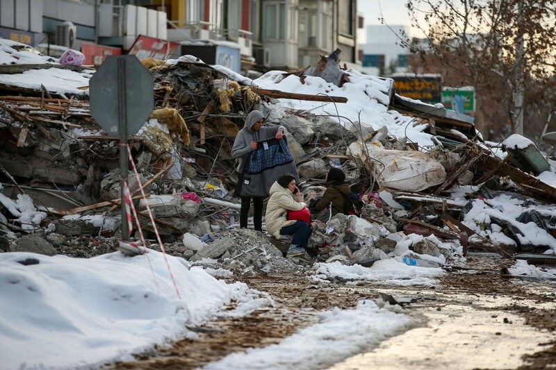 Menschen sitzen zwischen den Trümmern vor einem eingestürzten Gebäude. 