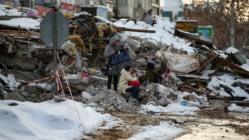 Menschen sitzen zwischen den Trümmern vor einem eingestürzten Gebäude.