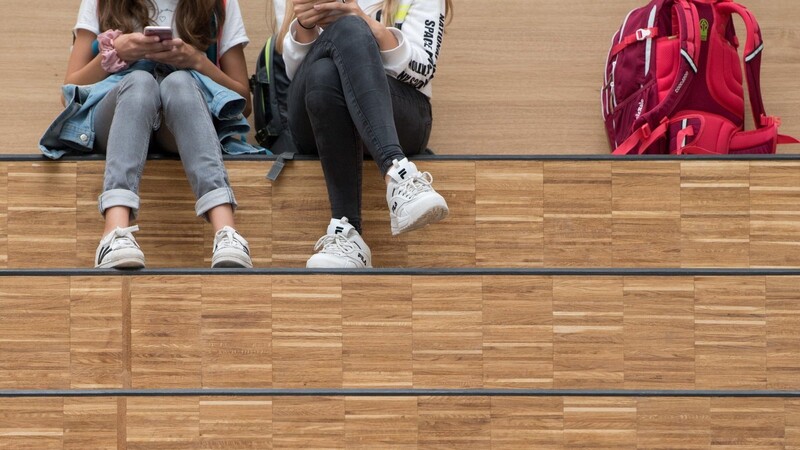 Schülerinnen sitzen im Foyer eines Gymnasium auf einer Freitreppe und halten jeweils ein Smartphone. In Schulen gelten eine Menge Corona-Regeln. Das könnte sich bald ändern.