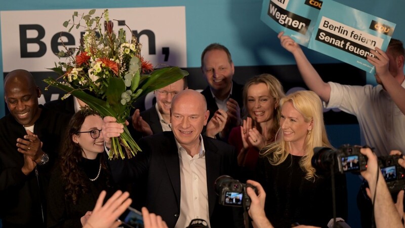CDU-Spitzenkandidat Kai Wegner (M.) steht mit einem Blumenstrauß in der Hand neben seiner Partnerin Kathleen Kantar (r.) auf der Bühne der Wahlparty seiner Partei.