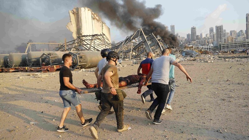 Helfer tragen ein Opfer der Explosion auf einer Trage, während im Hintergrund Rauch aufsteigt.