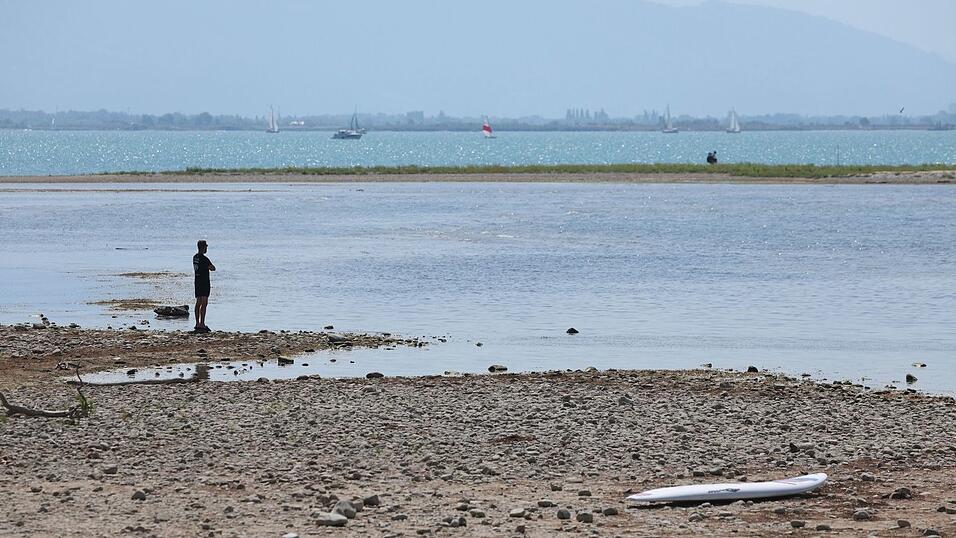 Es bleibt hei&szlig; in Bayern: Ein Mann steht am trockenen Ufer des Bodensees neben einem Surfboard. (Archivbild)
