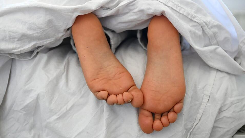 Die nackten F&uuml;&szlig;e und Pyjamahose einer Frau sind unter einer Bettdecke zu sehen.
