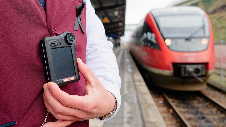 Ein Kundenbetreuer steht bei einem Pressetermin der Deutsche Bahn (DB) auf einem Bahnsteig neben einem Regionalzug und trägt eine Bodycam an der Dienstkleidung. Ein Kundenbetreuer steht bei einem Pressetermin der Deutsche Bahn (DB) auf einem Bahnsteig neben einem Regionalzug und trägt eine Bodycam an der Dienstkleidung.