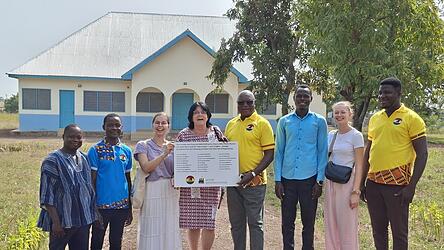 Vereinsmitglieder &uuml;bergeben die Spendertafel vor der neuen Krankenstation (im Hintergrund) an das Team des Schulzentrums. Mit den Lehrkr&auml;ften von links John Bosco, Stephen Tayo, Luise Altmann, Irmgard Hilmer, Schulleiter Pfarrer Dr. Dr. Camillo Bonsuuri, Idrussi Dawuda, Luisa Brunner und Joyous Joe Joseph.