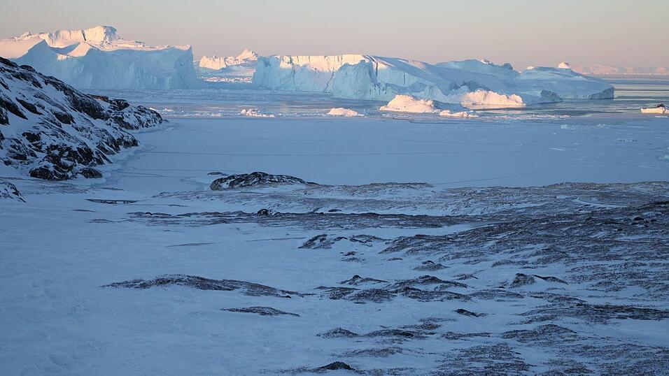 Eisberge ragen im Ilulissat-Eisfjord hinter einer verschneiten Gesteinslandschaft in die H&ouml;he.