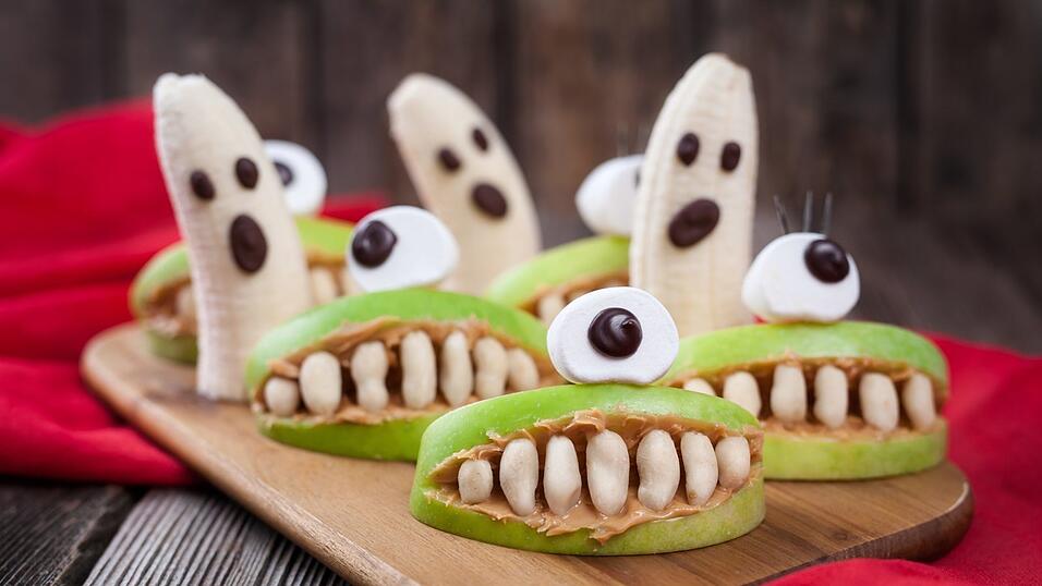 Ein gesunder Snack versteckt sich hinter diesen essbaren Monstern! Es sind gespenstische Zyklopenäpfel mit Nuss-Zähnen und einem Marshmallow-Auge. In der zweiten Reihe geistern Bananen umher.