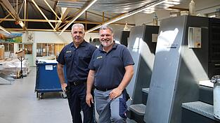 Christian Piller (links) und Thomas Pfeffer stehen vor einer F&uuml;nf-Farben-Offsetdruckmaschine. Damit kann man quasi alles drucken, sagt Pfeffer.