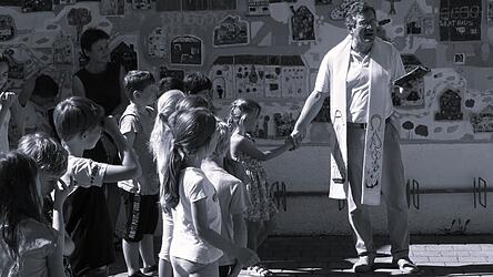 Pfarrer Alfons Dirscherl inmitten der Kinder mit Lehrerin Elfriede Bogner (links) bei der Einweihung des Wandfries an der Mauer der Grundschule, das im Schuljahr 2018/19 zusammen mit den beiden K&uuml;nstlern Georg Fuchssteiner und Peter Herr (Bogen) geschaffen wurde.